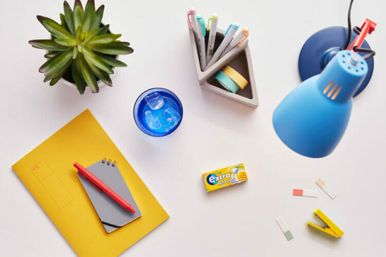 Picture shows a desk populated with a oencil case, lamp, notbooks, and a packet of Wrigley's Extra chewing gum.