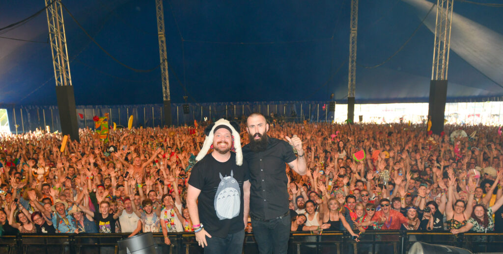 Dan Le Sac and Scroobius Pip stand beside each other on stage, with the crowd behind them, covered by a large blue tent. Taken at Bestival, Isle of Wight, in 2014, by Jules Annan.
